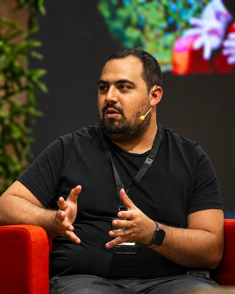 Michele Di Nardo speaking on stage, seated in a red armchair, wearing a headset microphone and an event badge.